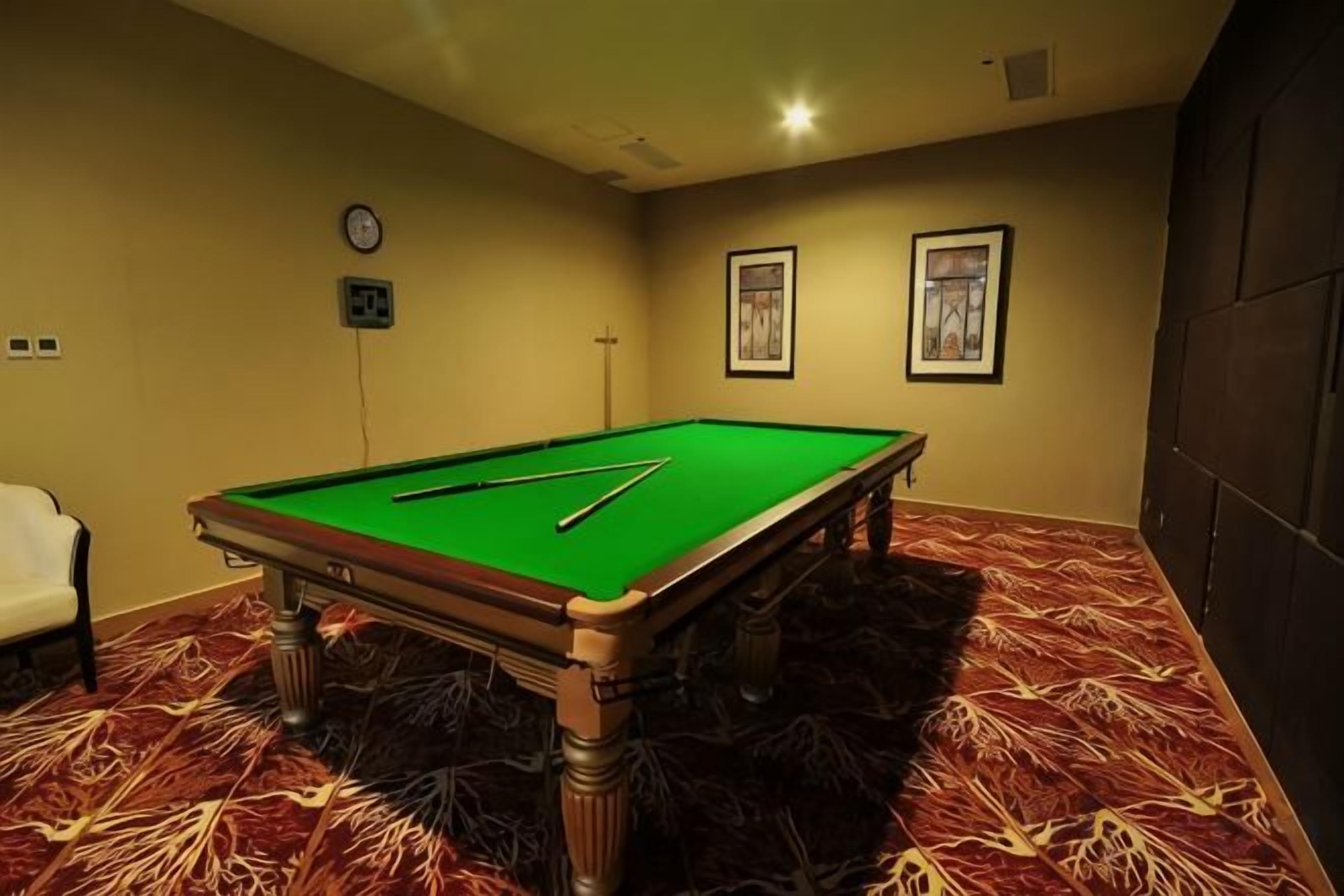 A pool table in a room with a patterned carpet, a clock on the wall, and framed pictures.
