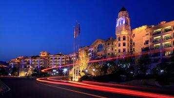 A hotel with a prominent clock tower, a flagpole, and a well-lit street with light trails.