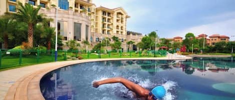 Una piscina con una persona buceando, rodeada de jardines con palmeras y un edificio grande al fondo.