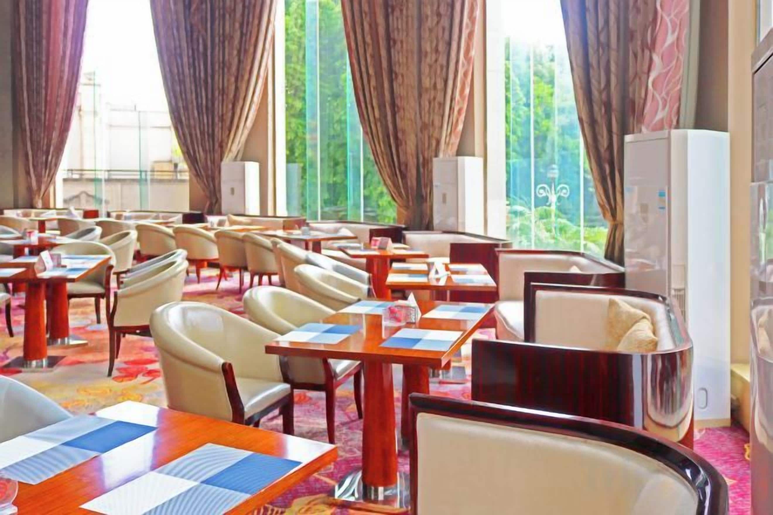 A hotel dining area with tables and chairs, large windows with curtains, and a refrigerator.