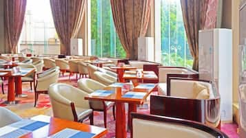 A hotel dining area with tables and chairs, large windows with curtains, and a refrigerator.