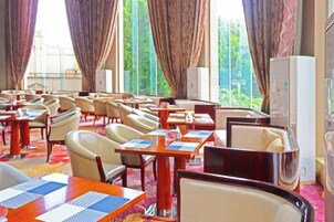 A hotel dining area with tables and chairs, large windows with curtains, and a refrigerator.