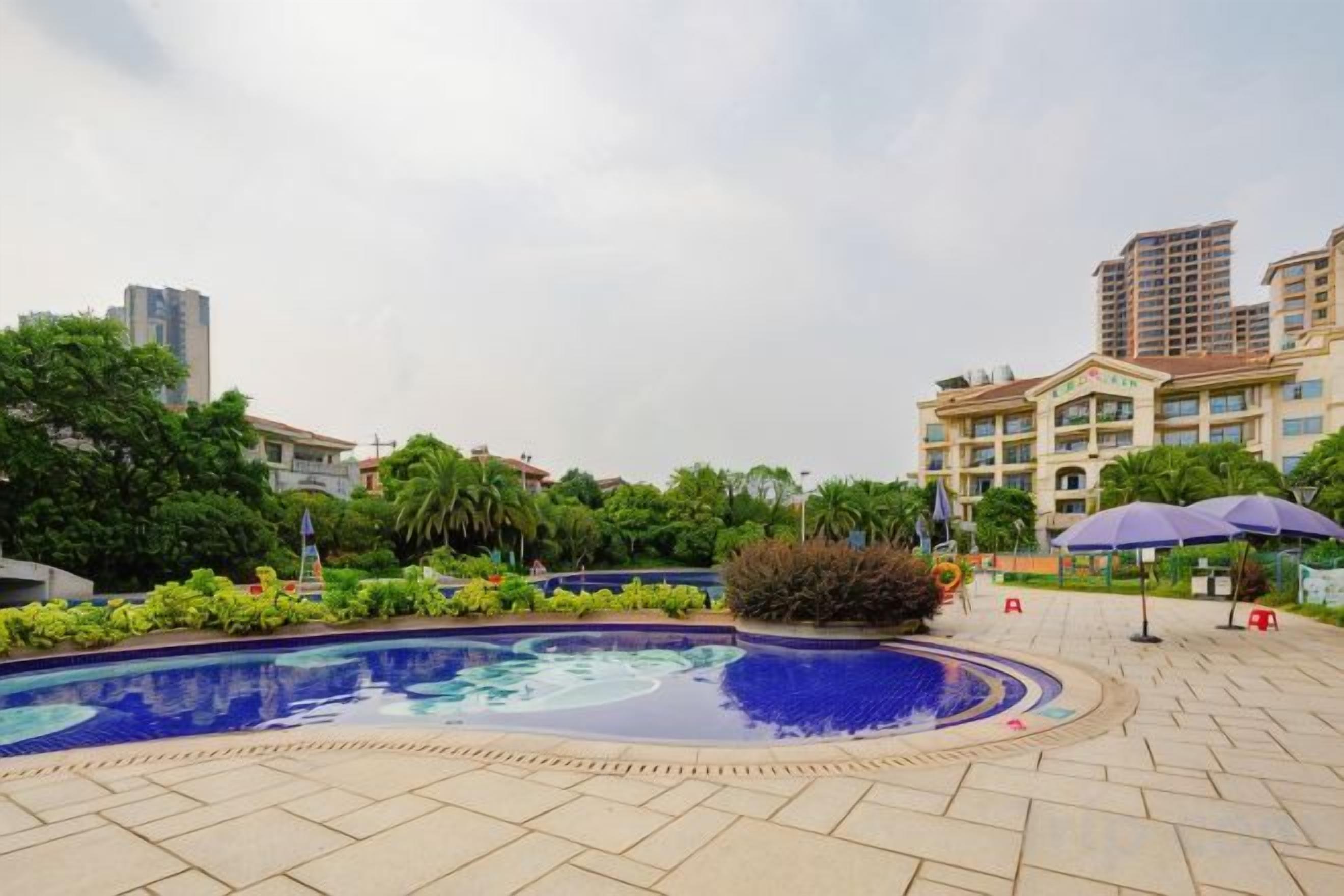 Une piscine dotée d’une terrasse carrelée, entourée de verdure et de bâtiments.