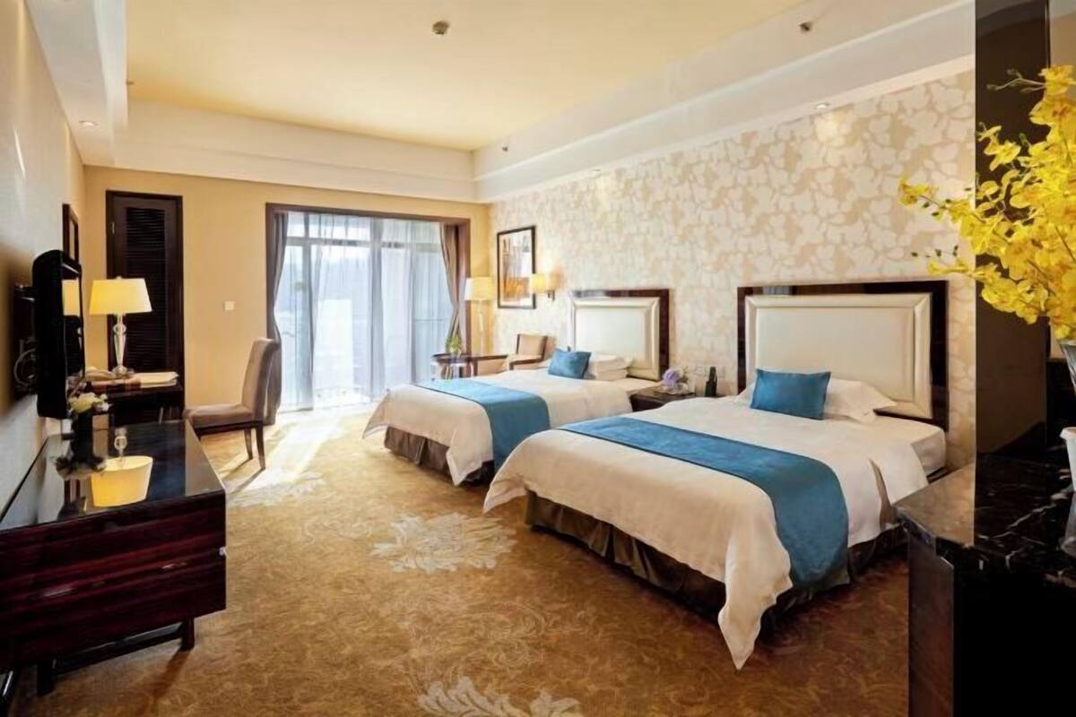A hotel room with two beds, a desk, a chair, a lamp, and a large window.