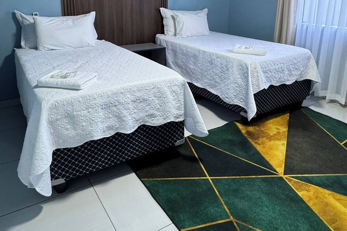 Une chambre d’hôtel avec deux lits simples, un mur lambrissé, une grande fenêtre avec des rideaux et un tapis à motifs.