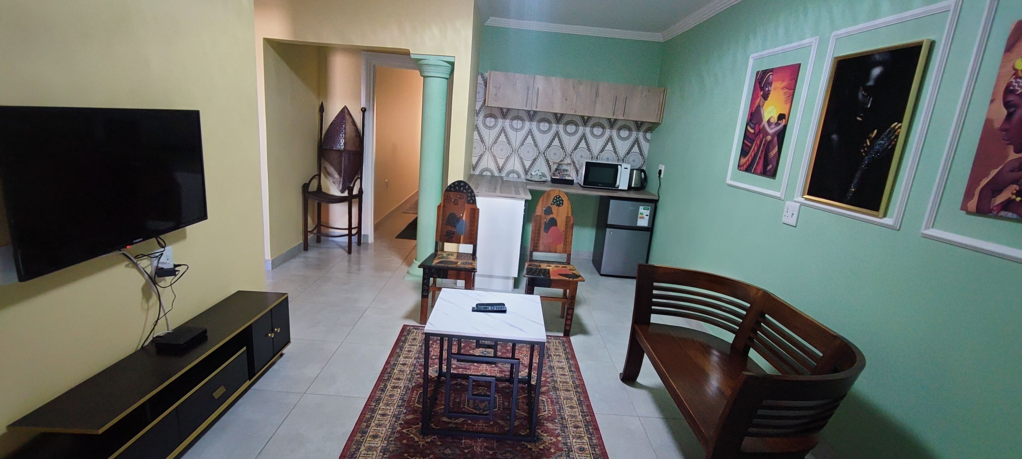 A living room with a flat-screen TV, a wooden bench, a small table, and a kitchen area with a microwave and refrigerator.