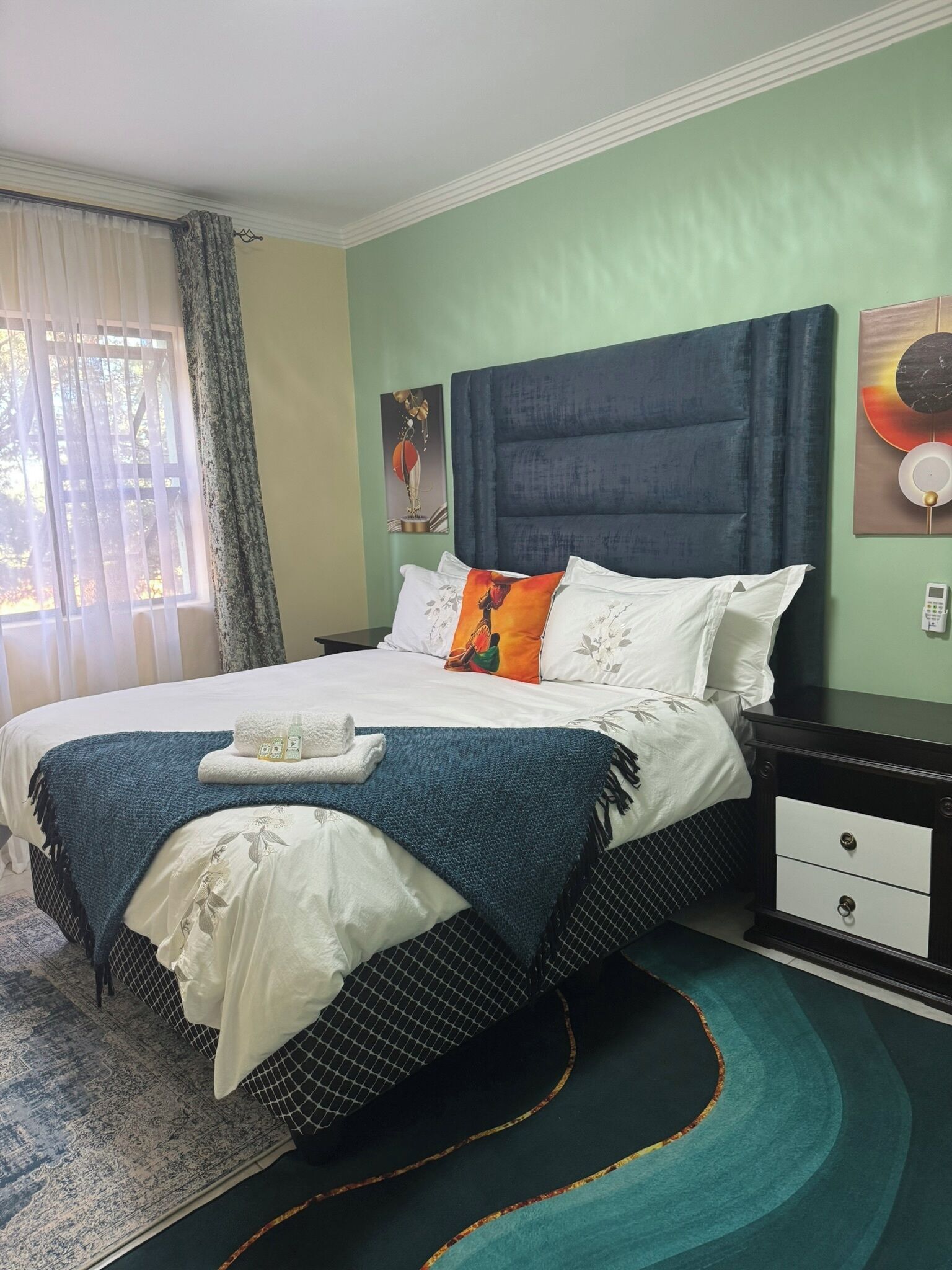 A neatly made bed with a blue headboard, white bedding, and decorative pillows. There is a bedside table with a phone and a small lamp. The room has a green wall and a window with curtains.