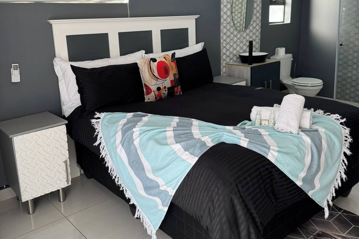 Une chambre moderne avec un grand lit, une table de chevet, une commode, une salle de bain avec des toilettes et un lavabo, et un mur décoré d’un motif.