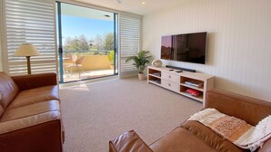 A living room with a large TV, two sofas, a ceiling fan, and a sliding glass door leading to a balcony. - Pool View, Lift Access 509, Opposite North Cronulla Beach (Cronulla)