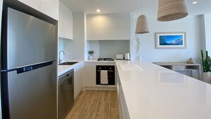 A modern kitchen with a stainless steel refrigerator, a dishwasher, a sink, and a stove. There are two hanging light fixtures and a framed picture on the wall. - Pool View, Lift Access 509, Opposite North Cronulla Beach (Cronulla)