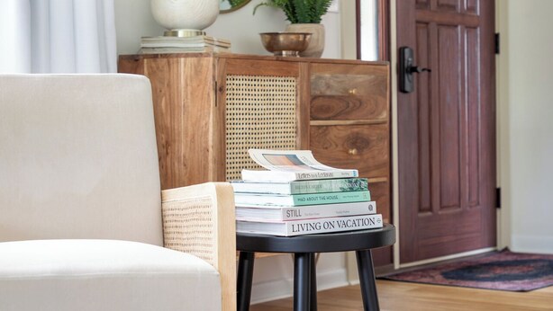 A cozy living room with a white sofa, a small wooden side table, and a stack of books on a black stool. A wicker chair and a woven pouf are also visible.