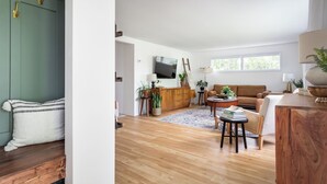 A spacious living room with a wooden floor, a sofa, a coffee table, and a TV mounted on the wall.