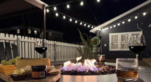 Un espace extérieur aménagé avec un foyer, une table où sont servis des plats et des boissons, et des guirlandes lumineuses.