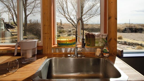 Une cuisine dotée d’un évier en acier inoxydable, de plans de travail en bois et offrant une vue sur un paysage naturel grâce à de grandes fenêtres.