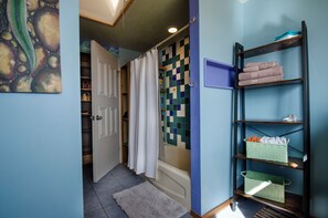 A bathroom with a bathtub, a shower curtain, a shelf with towels, and a wall with a mosaic tile design. - Earthship Stunning Views (Saguache County)