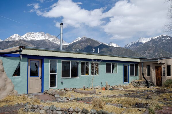 A single-story house with a blue exterior and a beige door, surrounded by a rocky landscape and mountains in the background. - Earthship Stunning Views (Saguache County)