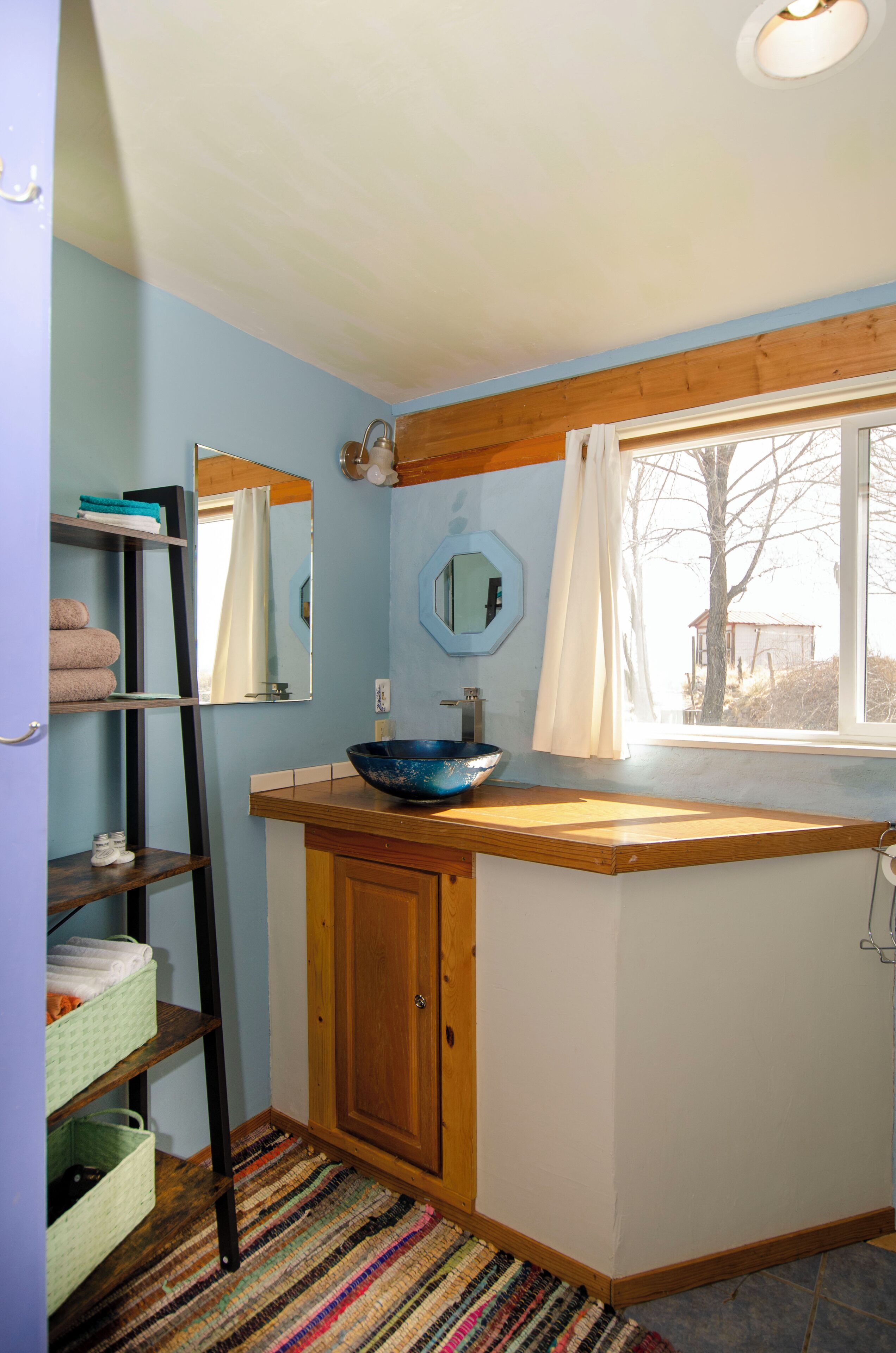 A compact bathroom with a wooden vanity, a blue basin, a mirror, and a window with a view of a tree.
