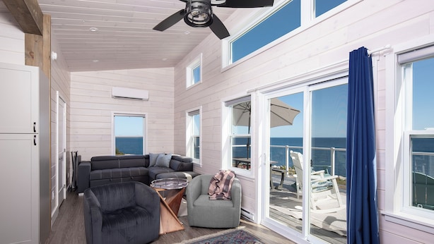 A modern interior with a ceiling fan, a sofa, a chair, and a view of the ocean through large windows.