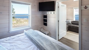 A bedroom with a bed, a TV mounted on the wall, and a view of the beach through a window.