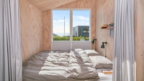 A bedroom with a large window, wooden walls, a bed with white bedding, and a small shelf with books and a hat. - Amazing home in Juelsminde (Juelsminde)
