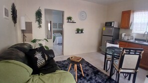 Una sala de estar con un sofá, una mesita redonda y un comedor con sillas. Hay una cocina con refrigerador, microondas y lavaplatos.