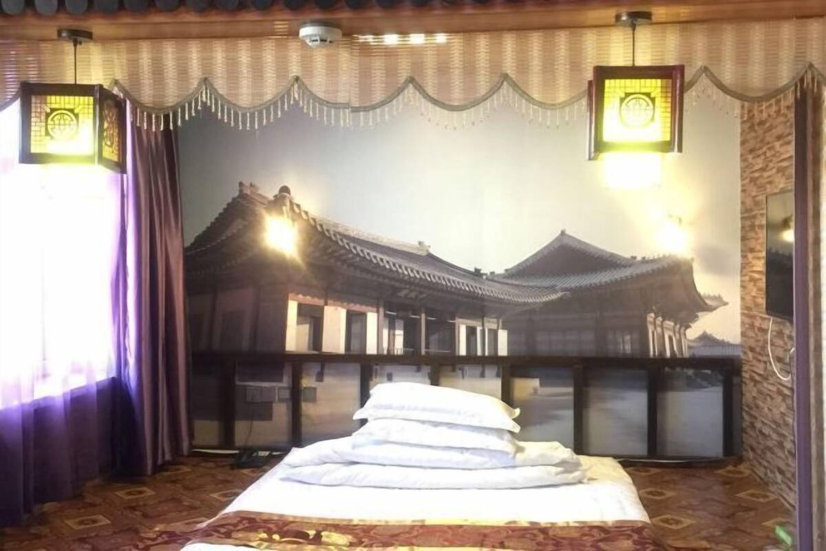 Une chambre ornée d’une fresque murale de style architectural chinois traditionnel, avec un lit recouvert de draps blancs et un tapis à motifs.