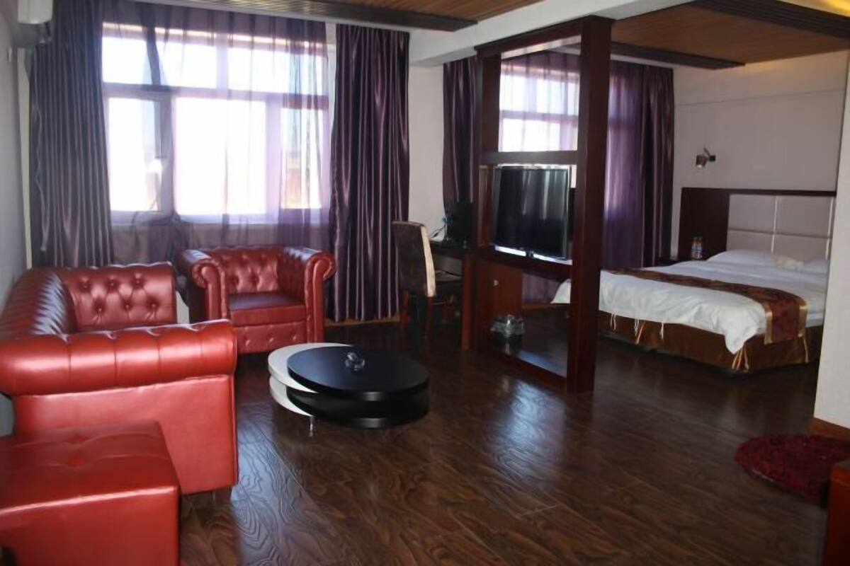 Une chambre d’hôtel avec un canapé en cuir rouge, un pouf rond, un lit avec une tête de lit blanche et un téléviseur.