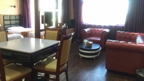 Een hotelkamer met een bed, een eettafel met stoelen, een rode leren bank, een klein rond tafeltje met een lamp en een groot raam met gordijnen.