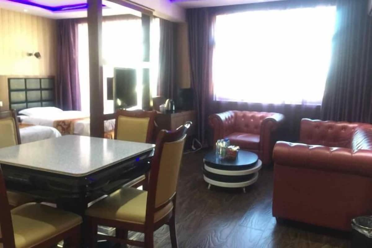 Une chambre d’hôtel comprenant un lit, une table à manger avec des chaises, un canapé en cuir rouge, une petite table ronde avec une lampe, et une grande fenêtre avec des rideaux.