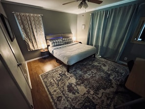 Une chambre à coucher comprenant un lit, une table de chevet, un ventilateur de plafond, un tapis à motifs et une fenêtre avec des rideaux.
