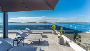 Un balcon agrémenté de chaises longues, d’une table et de plantes en pot, donnant sur une étendue d’eau où voguent des voiliers.