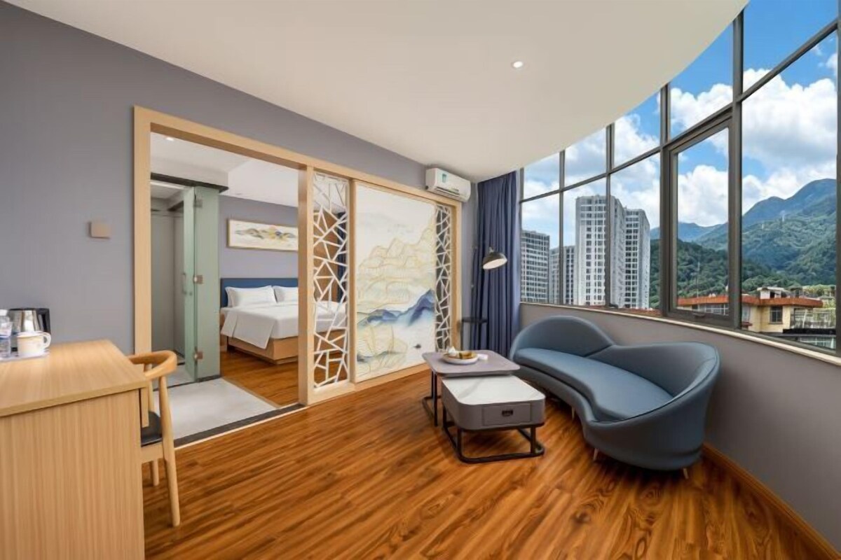 A hotel room with a wooden floor, a bed, a desk, a chair, and a view of mountains through large windows.