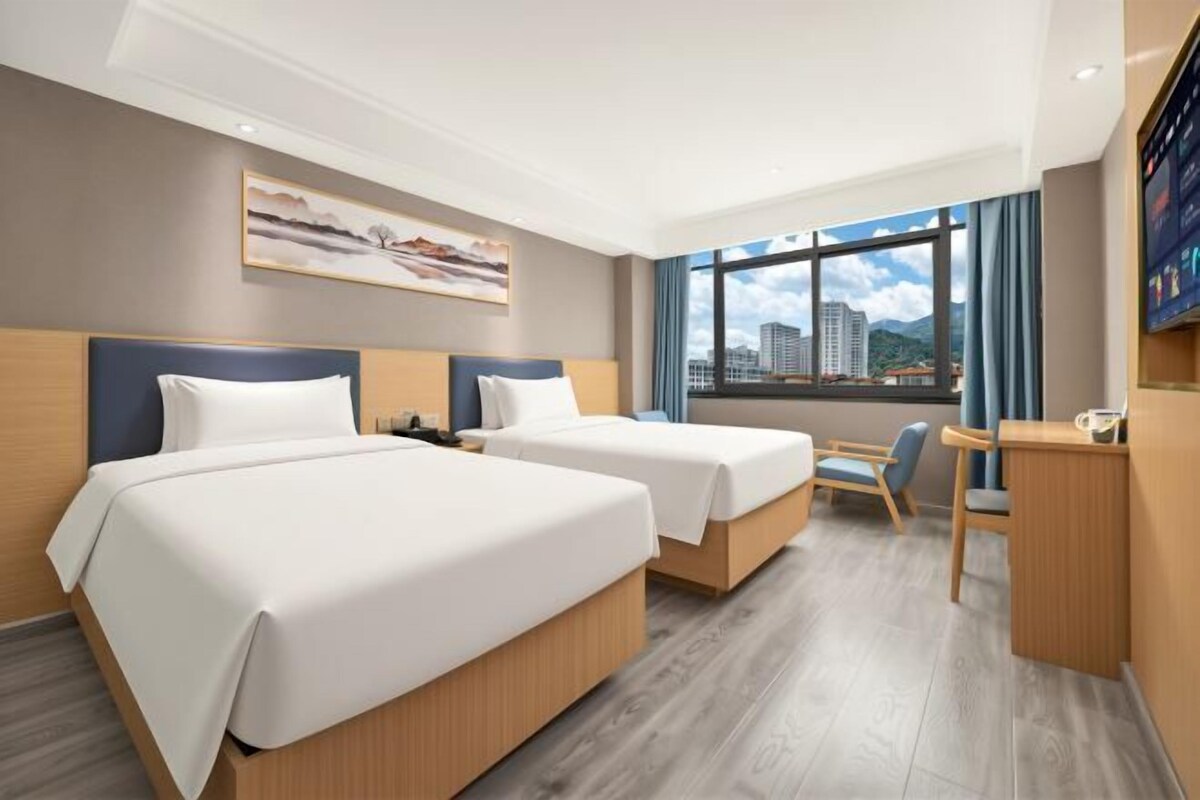 A hotel room with two beds, a desk, a chair, and a large window with a city view.