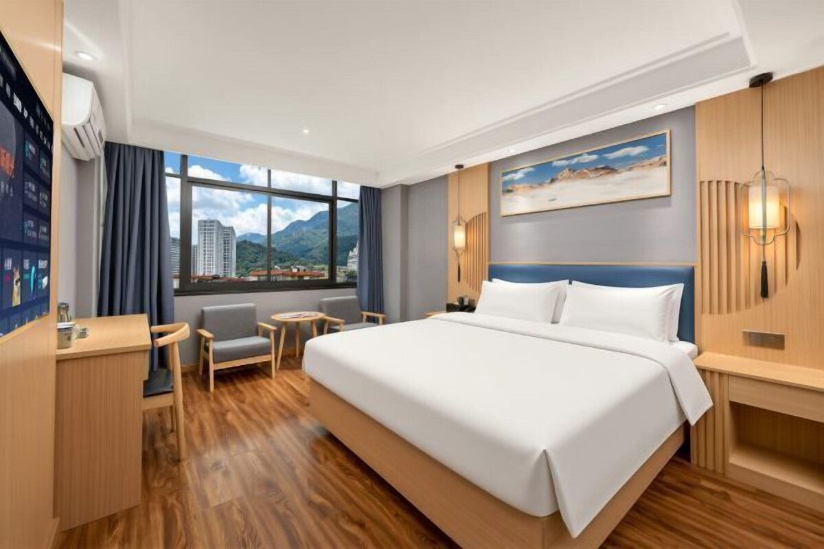A hotel room with a large bed, a desk, a chair, a small table, and a view of a cityscape through the window.