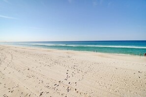A wide sandy beach with footprints leading to the water, a clear sky, and a few people in the distance.