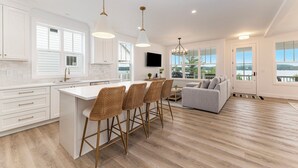 A modern kitchen with white cabinets, a large island, and bar stools. There is a dining area with a gray sofa and a TV mounted on the wall. - Luxe Lakefront Living on Stunning Crystal Lake in Benzonia, MI (Benzonia)