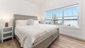 A bedroom with a large window, a wooden headboard, a white bedspread, and a bedside table with a lamp. - Luxe Lakefront Living on Stunning Crystal Lake in Benzonia, MI (Benzonia)