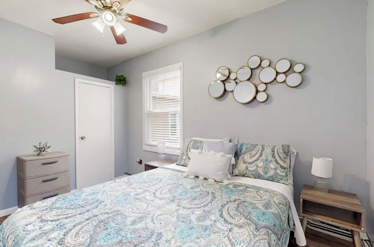 Une chambre à coucher comprenant un lit, un ventilateur de plafond, un miroir fixé au mur, une commode et une fenêtre avec des stores.