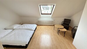 A small, bright room with a sloped ceiling, a skylight, a bed with white bedding, a radiator, a small round table, and a chair. - City Apartment - Business, Uni, Handwerker - Free Wifi (Bochum)