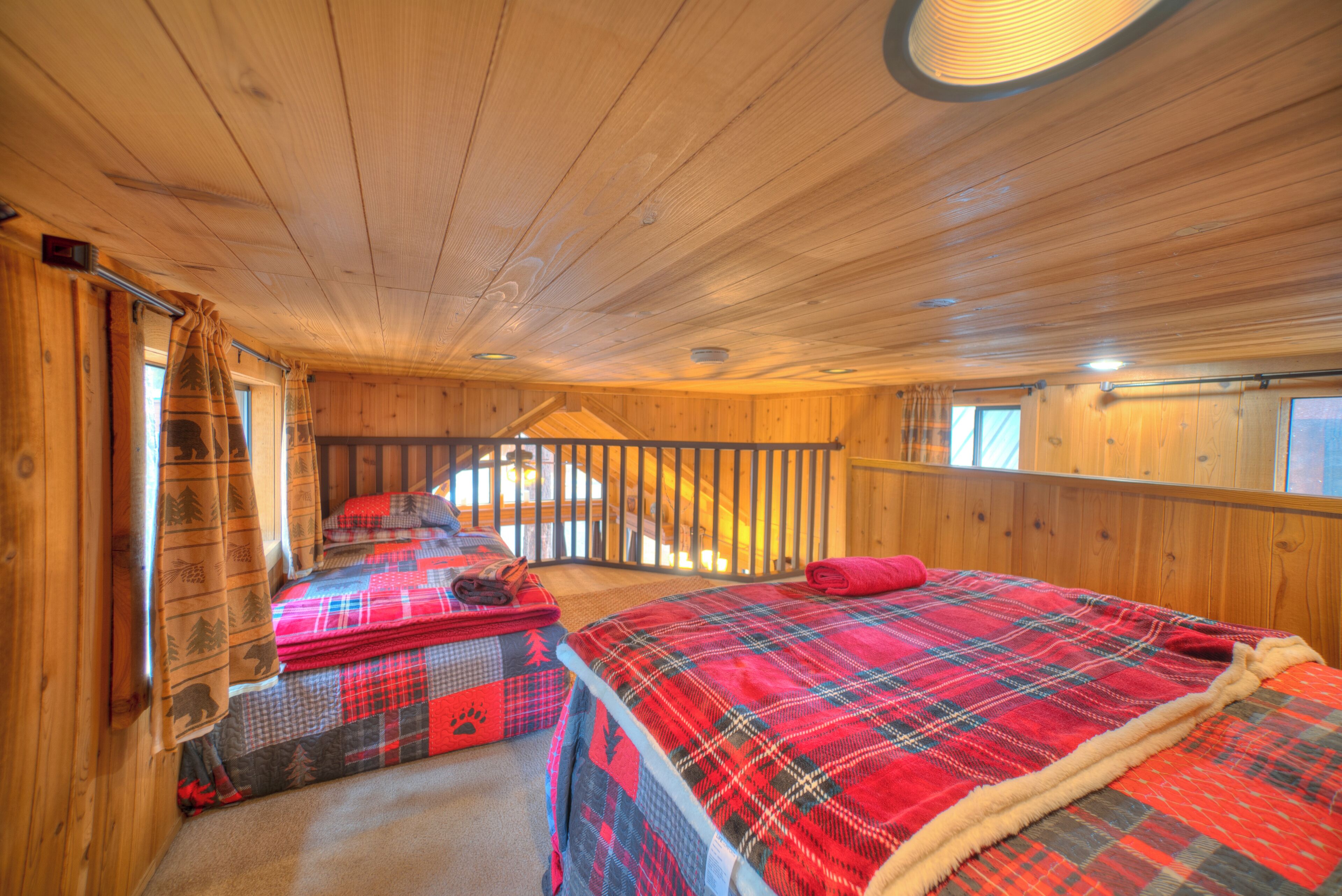A cabin interior with wooden walls and floors, a bed with a plaid blanket, a window with a view, and a staircase leading to an upper level.
