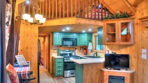 Interior de una cabaña de madera con cocina, que incluye gabinetes verdes, microondas, estufa y televisor.