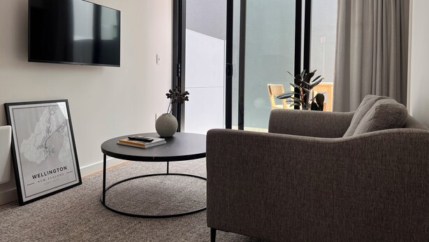 A modern living room with a flat-screen TV, a round coffee table with a vase and books, a grey sofa, and a large window with curtains.