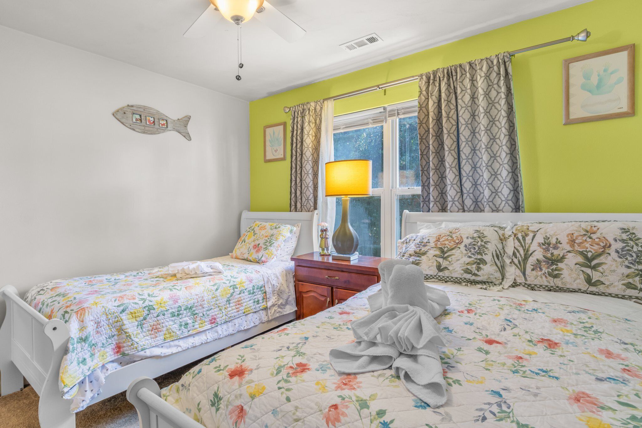 A bedroom with two single beds, a window with patterned curtains, a fish decoration on the wall, and a lamp on the nightstand.