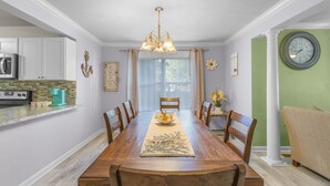 Une salle à manger avec une table en bois, des chaises, un lustre et un coin cuisine équipé de placards et d’appareils électroménagers.