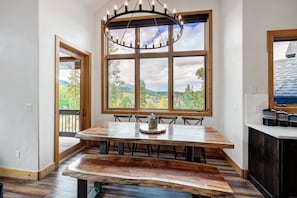 Un espace repas avec une grande table en bois, un lustre et une vue sur un paysage montagneux, visible à travers de grandes fenêtres.