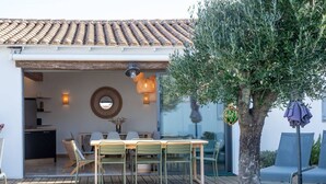 An outdoor dining area with a wooden table and chairs, a hanging light fixture, and a view of the kitchen through a glass door. - Alma - Renovated villa with heated pool few blocks from the beach (LE BOIS PLAGE EN RE)