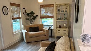Una sala de estar acogedora con un sofá color beige, una mesita auxiliar pequeña y un ventilador de techo. Hay dos ventanas con persianas, una planta y un estante con objetos decorativos.