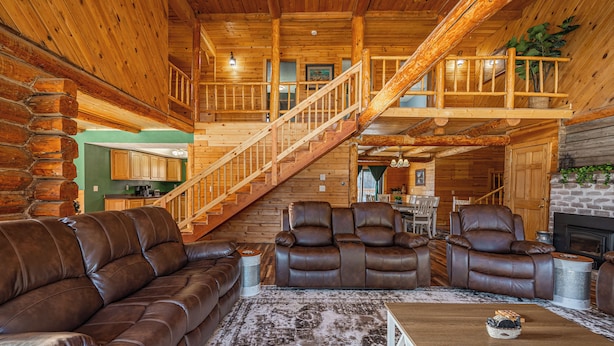 Interior de una cabaña de troncos con un sofá de cuero, dos sillones reclinables, una mesa de centro de madera y una escalera que lleva a un nivel superior.