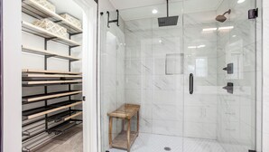 Ein begehbarer Kleiderschrank mit Regalen und einer Holzbank, eine Duschkabine mit Glas und einer Handbrause sowie ein kleiner hölzerner Hocker.