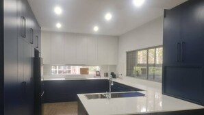 A modern kitchen with dark cabinetry, a central island with a sink, and recessed ceiling lights. - Sanctuary on the Creek (Sanctuary Point)